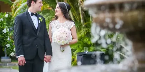 A bride and groom standing together in a tropical garden with a fountain in the foreground.