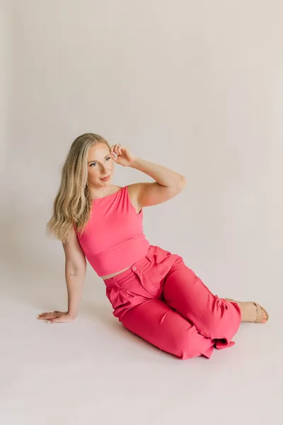 A woman with styled hair and makeup sitting on a studio floor wearing a pink top and pants.