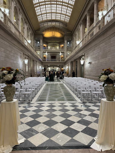 A grand hall interior set up for an event with rows of white folding chairs and draped tables.