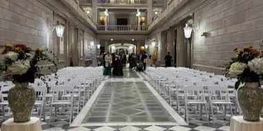 A grand hall interior set up for an event with rows of white folding chairs and draped tables.