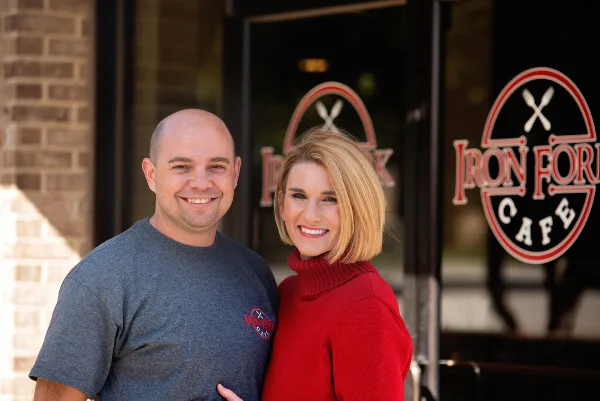 Two smiling owners standing in front of the Iron Fork Cafe storefront with the business logo visible on the window.