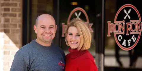 Two smiling owners standing in front of the Iron Fork Cafe storefront with the business logo visible on the window.