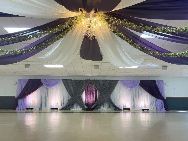 A wedding venue interior featuring purple and white ceiling draping, a crystal chandelier, and a decorated stage backdrop with grey and lavender linens.