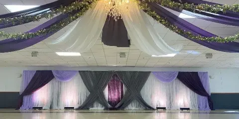 A wedding venue interior featuring purple and white ceiling draping, a crystal chandelier, and a decorated stage backdrop with grey and lavender linens.
