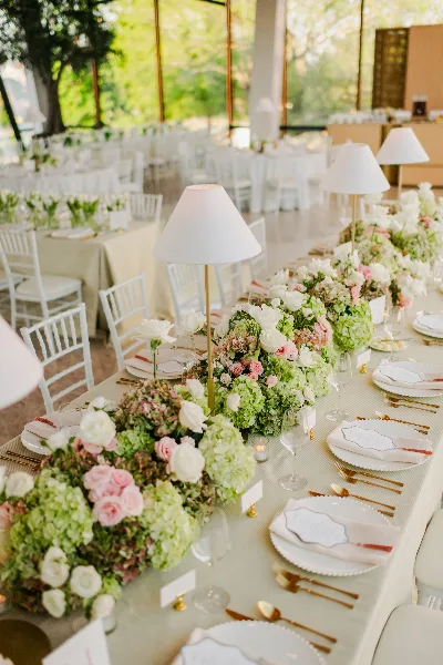 A long banquet table set for a wedding reception featuring lush floral arrangements, white table lamps, and elegant place settings.