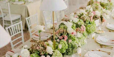 A long banquet table set for a wedding reception featuring lush floral arrangements, white table lamps, and elegant place settings.