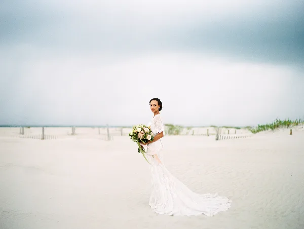 A bride in a white lace gown holding a bouquet, standing on a sandy beach with a cloudy sky in the background.