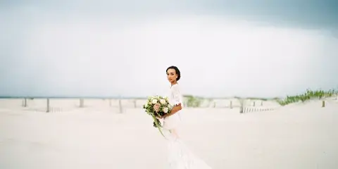 A bride in a white lace gown holding a bouquet, standing on a sandy beach with a cloudy sky in the background.