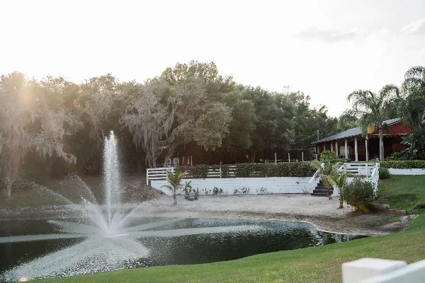 Outdoor event venue featuring a pond with a fountain, white fencing, and a rustic pavilion under a bright sky.