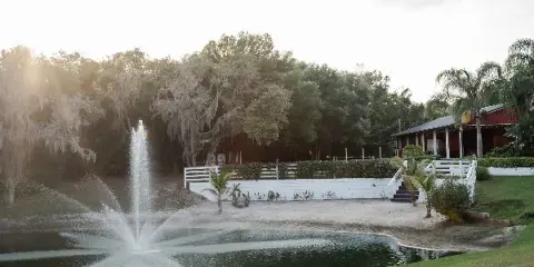 Outdoor event venue featuring a pond with a fountain, white fencing, and a rustic pavilion under a bright sky.