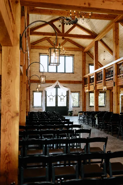 Interior view of a rustic timber-frame wedding barn with rows of black chairs and a chandelier.