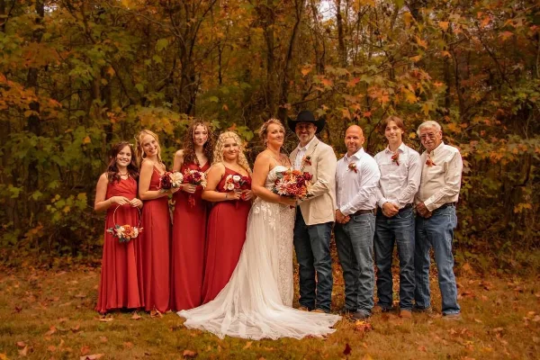A wedding party posing outdoors in a wooded area with fall foliage.