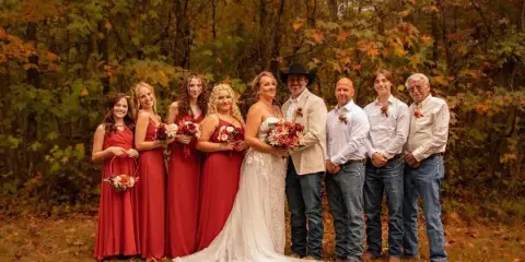 A wedding party posing outdoors in a wooded area with fall foliage.