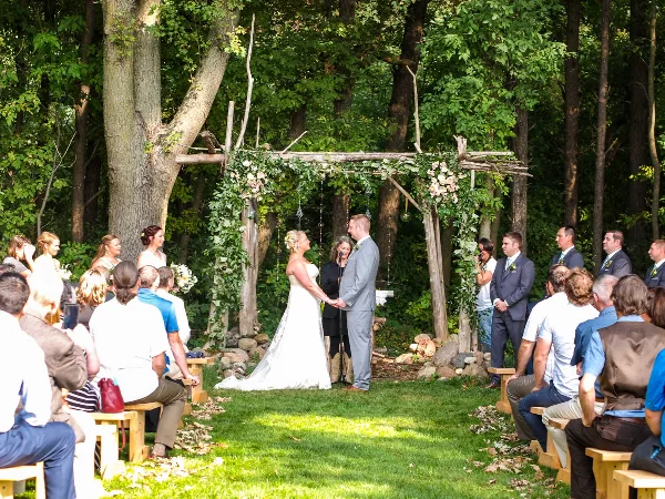 A bride and groom exchanging vows under a rustic wooden arch decorated with greenery and flowers in a forest clearing, surrounded by seated guests.