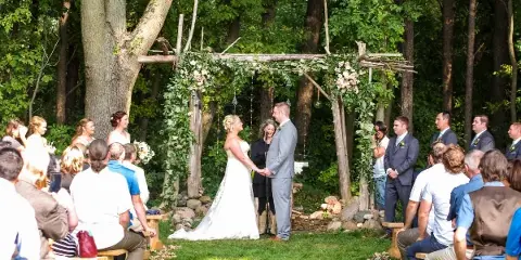 A bride and groom exchanging vows under a rustic wooden arch decorated with greenery and flowers in a forest clearing, surrounded by seated guests.