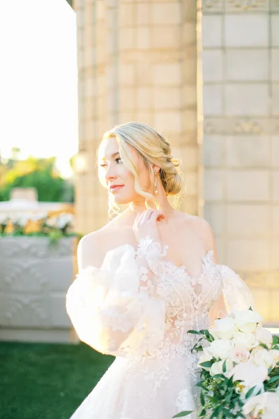 A bride with styled blonde hair and makeup wearing a white lace wedding dress and holding a bouquet of white roses.