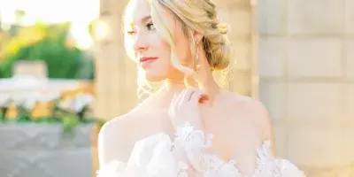 A bride with styled blonde hair and makeup wearing a white lace wedding dress and holding a bouquet of white roses.