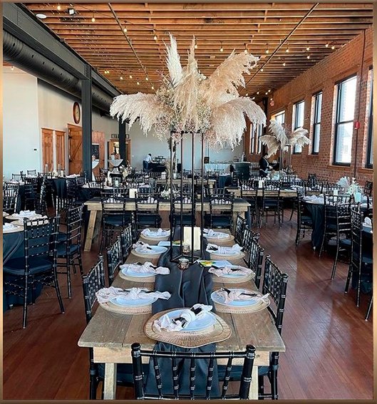 Interior view of a modern-industrial event space with exposed brick walls, wood floors, and set dining tables with black chairs and linens.