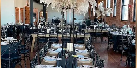 Interior view of a modern-industrial event space with exposed brick walls, wood floors, and set dining tables with black chairs and linens.