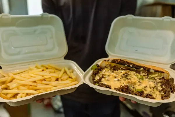 A person holding two white catering containers, one containing a cheesesteak with melted cheese and the other containing french fries.