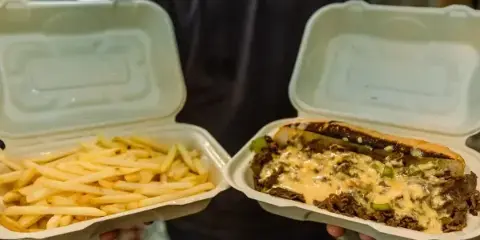 A person holding two white catering containers, one containing a cheesesteak with melted cheese and the other containing french fries.