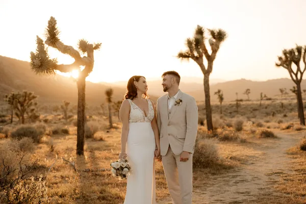 A bride and groom standing together in a desert landscape with Joshua trees at sunset.