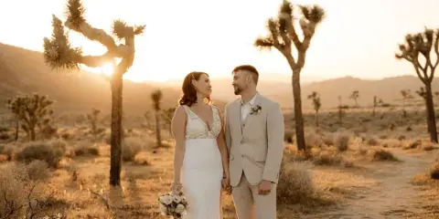 A bride and groom standing together in a desert landscape with Joshua trees at sunset.
