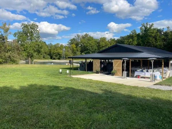 A wide shot of a large open-air wedding pavilion with a dark metal roof, situated on a green lawn with a pond and trees in the background.