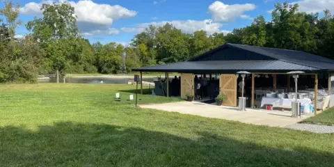 A wide shot of a large open-air wedding pavilion with a dark metal roof, situated on a green lawn with a pond and trees in the background.