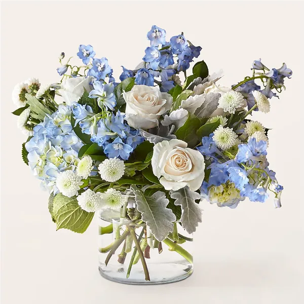 A floral arrangement featuring white roses, blue delphiniums, and white chrysanthemums in a clear glass vase.