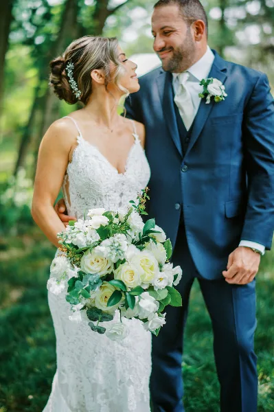 A bride with a styled updo and makeup standing next to a groom in a blue suit outdoors.