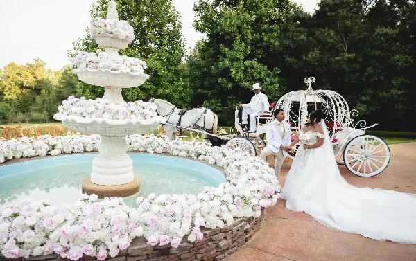A bride and groom standing near a white horse-drawn carriage and a large floral fountain at an outdoor wedding venue.