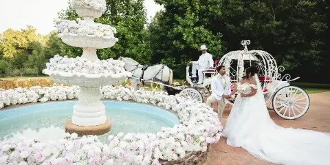 A bride and groom standing near a white horse-drawn carriage and a large floral fountain at an outdoor wedding venue.
