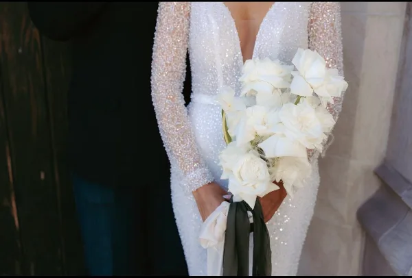 A bride in a white beaded gown holding a bouquet of white roses and calla lilies.