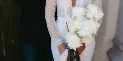 A bride in a white beaded gown holding a bouquet of white roses and calla lilies.