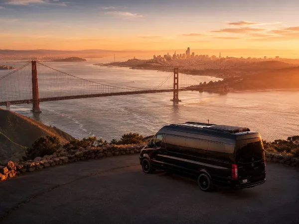 A black luxury Sprinter van parked on a scenic overlook with the Golden Gate Bridge and San Francisco skyline in the background at sunset.