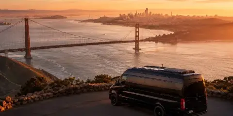 A black luxury Sprinter van parked on a scenic overlook with the Golden Gate Bridge and San Francisco skyline in the background at sunset.