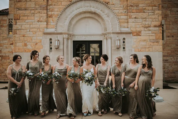 A bride and her bridesmaids in olive green dresses standing in front of a stone church.