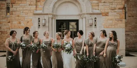 A bride and her bridesmaids in olive green dresses standing in front of a stone church.