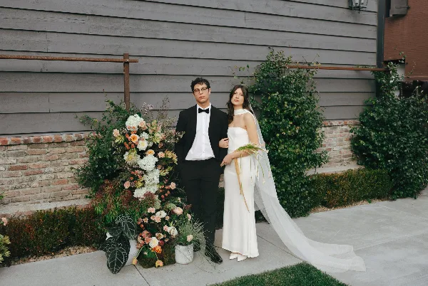 A bride and groom in formal wedding attire standing together next to a large floral arrangement outdoors.