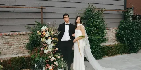 A bride and groom in formal wedding attire standing together next to a large floral arrangement outdoors.