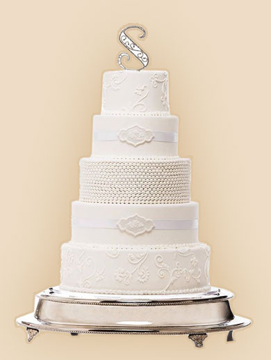 A five-tier white wedding cake with textured and piped decorations, a white ribbon, and a silver 'S' topper on a silver cake stand.