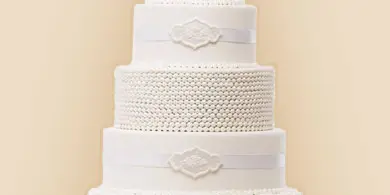A five-tier white wedding cake with textured and piped decorations, a white ribbon, and a silver 'S' topper on a silver cake stand.