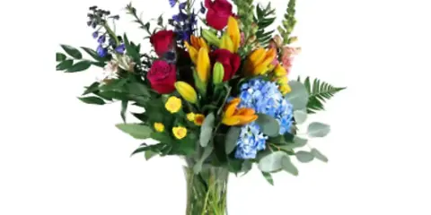 A mixed floral bouquet containing red roses, yellow lilies, blue hydrangeas, and greenery arranged in a clear glass vase.