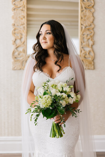 A bride in a white wedding dress and veil holding a bouquet of white flowers, showcasing styled hair and makeup.