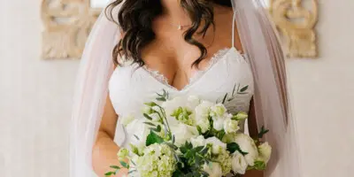 A bride in a white wedding dress and veil holding a bouquet of white flowers, showcasing styled hair and makeup.