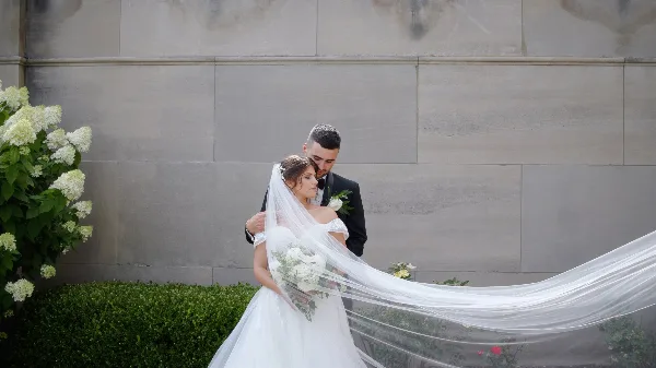 Bride and groom embracing outdoors with a long flowing veil and white bouquet against a stone wall.