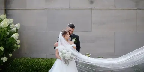 Bride and groom embracing outdoors with a long flowing veil and white bouquet against a stone wall.