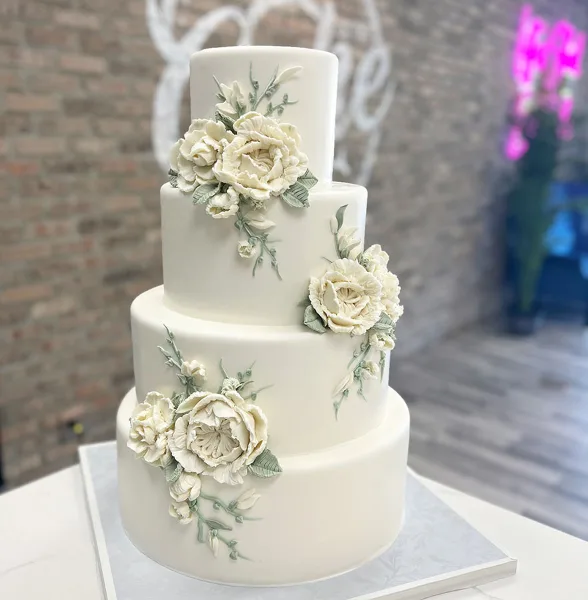 A four-tier white wedding cake decorated with white sculpted flowers and green leaves, set against a brick wall background.