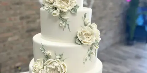 A four-tier white wedding cake decorated with white sculpted flowers and green leaves, set against a brick wall background.